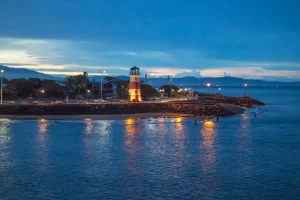 Lighthouse on a rocky coast at night, with mountains in the background and the water reflecting the lighthouse's bright orange light.